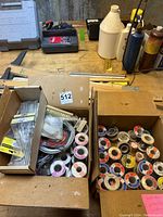Two cardboard boxes on a wooden workbench containing multiple solder spools and packaged acid brushes