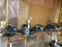 Multiple electric soldering irons and controllers set on a wood shelf against pegboard and plastic sheeting. Cords are tangled. Various soldering iron stands present.