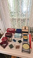 Overview of twelve stained glass decorative boxes and coaster holders in different colors and shapes on white table near lace curtain.