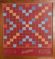 Full view of deluxe Scrabble game board with burgundy and multi-color classic scoring squares, gold trim, and logo.