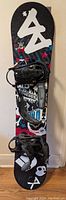Photo shows the full length of the child's snowboard standing vertically against a wall. The snowboard has a colorful graphic design with stars, skulls and various patterns. Two black Indy brand bindings are attached.