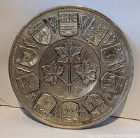 Front view of the round vintage brass Canada plate showing embossed provincial crests, central maple leaf motif, and textured background.