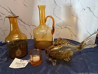 Four amber glass items displayed on a cloth including two jugs, a fish sculpture, and a candle holder.