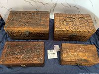 Top view of four rectangular wooden chests with various Asian hand carved depictions including boats, figures, and floral leaf details.
