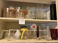 Wide shot showing a shelf with multiple glass vases of various shapes and colors including clear, white floral, yellow ribboned, dark slender, and red glass vases.