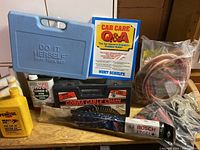 Full display of lot items on a wooden surface including oil bottles, tool case, book, Bosch wiper, jumper cables in bags, and Cobra cable chains box.