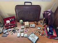 Full overview of the collection on wooden surface showing leather briefcase, dopp kit, pins, medals, tie clips, keys, watches in boxes, and decorative stones.
