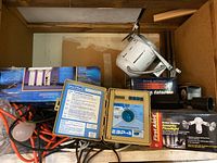 Photo showing the full lot contents including boxes of shop lights, faucet water filtration system, Rainbird ESP-4 sprinkler timer open with visible programming dial, motion detector security lights in box, and electrical wiring tangled and bagged.