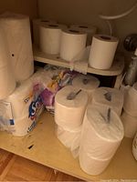 Photo showing several loose individual toilet paper rolls on a cabinet shelf next to a sealed Scott pack and large plastic wrapped toilet paper rolls.