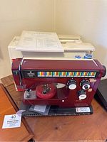 Front side view of burgundy Viking Selectronic 6570 vintage sewing machine on wooden table with instruction manual on top and red pincushion with pins attached.