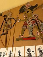Two vintage Southeast Asian shadow puppets mounted on a cardboard backing showing front and side views, highlighting their intricate painted details and articulated limbs with rare lucite or bakelite handles.