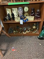 Two wooden shelves filled with assorted decorative items: anniversary clock, marble bookends, figurines, black carved chairs, alabaster boxes, brass decor pieces.