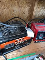 Two battery chargers side by side on a wooden shelf in a garage, showing full units with power cords and connection cables.