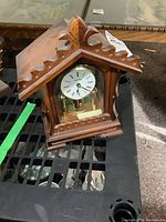 Front view of wooden anniversary style clock with round white clock face, Roman numerals and glass sides revealing spinning dancers inside.