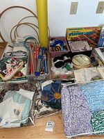 Overview of crafting supplies including wooden hoops, knitting needles, plastic containers with beads, lace trim, yarn bundles, and fabric pieces piled on the floor around the weaving loom box.