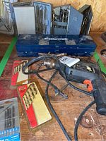 View of the Black & Decker electric drill with Mastercraft blue metal drill bit case and several additional drill bit cases scattered on a wooden surface