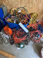 Blue plastic tote loaded with assorted extension cords and cables in various colors and lengths, along with power strips and a trouble light partially visible.