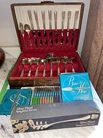 Photo of open wooden canteen with red velvet lining holding silver plated flatware including forks, knives, spoons, and a collectible spoon set along with boxed stainless steel knives and a silver plated spaghetti server.