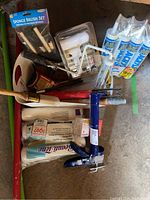 Full lot view showing acrylic caulk tubes, paint brushes, sponge brush set, caulking tool, paint rollers, and roller trays on floor.