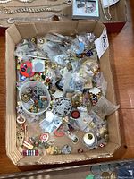 Photo shows a cardboard box filled with various pins and badges, some in individual plastic bags and some loose, including colorful and metallic pieces