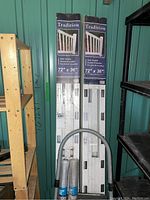 Two unopened boxed white vinyl stair railing sections, 72" x 36" each, stacked upright against a teal wall with shelving on each side. Metal step stool with curved handle and replacement screen rolls are in front.