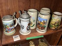 Photo showing all six beer steins displayed on a wooden shelf. Two steins have lids, each stein is distinct in decoration and color.