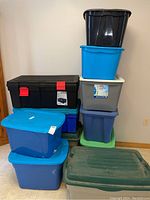 Stacked eleven plastic storage totes in different colors and sizes against a wall, showing general condition and quantity