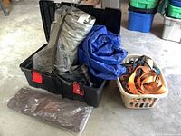 Black plastic storage tote containing gray and blue tarps, with an orange tag on the latch, photographed against a concrete floor.