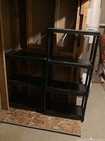 Two black stackable plastic utility shelves with three tiers each, placed in a basement or storage area with unfinished walls.