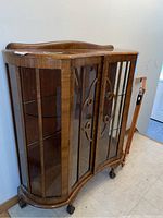 Front angled view of wooden display cabinet with curved glass and carved front doors, on casters