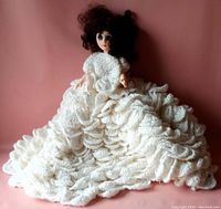 Bride doll with black curly black hair seated wearing a voluminous white hand-crocheted dress with scalloped trim against a pink background.