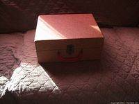 Closed suitcase style record player with brown textured casing, red handle, and metal latch on a quilted surface with sunlight casting shadows.
