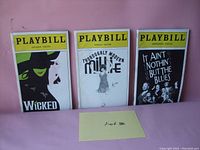 Photo showing three vintage Playbills from Wicked, Thoroughly Modern Millie, and It Ain't Nothin But the Blues on a pink background with a lot number card.