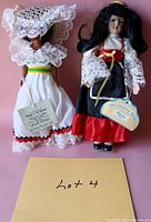 Front view of two international bride dolls, one in white lace bonnet and dress, the other in red and black outfit with tag.