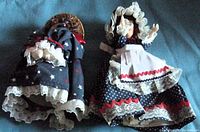 Two cloth dolls in patriotic red, white, and blue fabrics standing on surface. One doll has bonnet and apron, the other doll has cone-shaped bottom.