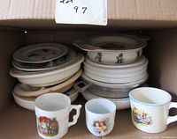 Photo showing three small antique baby cups in front and a stack of various small baby dishes and plates behind them inside a cardboard box.