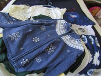 Blue zip-up sweater with white star/snowflake embellishments and a mix of white and colored women's clothes beneath it, showing detail of sweaters and fabric textures.