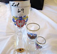 Three vintage German glass items with gold trim and decorative painted designs, including a tall glass marked Frankfurt A.M. and two smaller glasses with German motifs, displayed on white fabric.