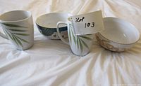 Photo showing two white ceramic mugs with green leaf prints, one ceramic bowl with a blue and white design, and one ceramic bowl with beige and white design.