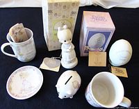A shot showing multiple Precious Moments items including figurines, mugs, plate, piggy bank and original boxes all arranged on a black surface.