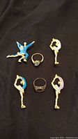 Top view of jewelry items arranged on black background, showing dancer figurine pins and two rings.