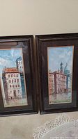 Photo showing two vertical rectangular paintings side-by-side, in dark brown frames depicting Venice canals and buildings.
