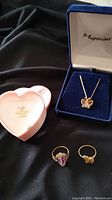 Three pieces laid out on black cloth: pink heart jewelry box, blue velvet box with butterfly necklace, two gold rings
