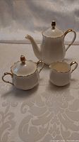 Photo of white porcelain tea pot, sugar bowl, and creamer all with gold rim accents set on a patterned tablecloth.