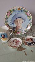 Large decorative plate with floral border and portrait of Queen Mum, standing in background the cups and pin dishes on a fabric surface