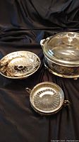 Three items including a clear glass Pyrex covered dish with lid, one round silver plate tray with decorative cut out design, and one smaller oval silver plate tray with gold tone handles shown against black fabric background.