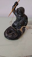 Side view of black soapstone carving of fisherman holding fishing rod with string attached, seated on shaped base with two fish carved in relief.