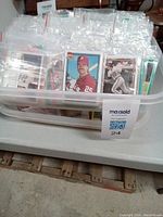 Small clear plastic storage bin filled with baseball cards in individual plastic sleeves, upright and organized. Cards exhibit various players and teams.