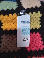 Close-up photo showing the colorful crocheted squares with scalloped edges and a MaxSold lot number tag attached to the blanket.