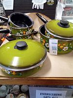 Photo showing several green enamel pots and pans with black handles and colorful floral and fruit designs on an olive green background. Includes lids, though some lids or handles are missing.